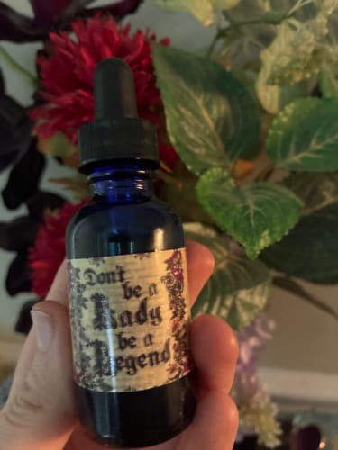 Customer's hand holding the small blue dropper bottle of Don't Be A Lady, Be A Legend Hair and Body Oil in front of flowers.