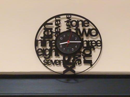 Ein Rezensent zeigt seine schwarze Vinyl-Wanduhr mit einem Zifferblatt aus Wörtern an einer Wand.