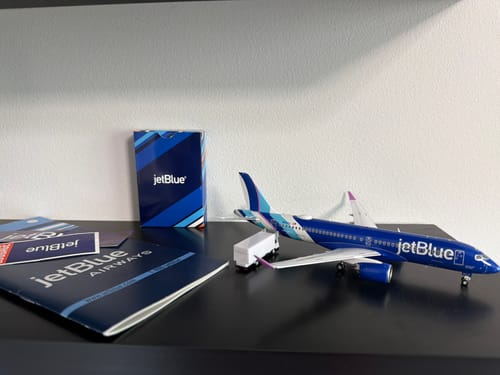 Customer's blue Gemini200 JetBlue A220-300 model airplane displayed on a dark shelf with other branded items.
