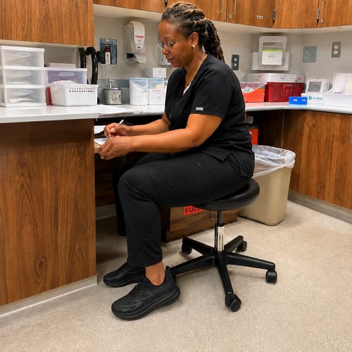 Customer wearing black Ortho Walk™ shoes while sitting on a stool in a medical office setting.