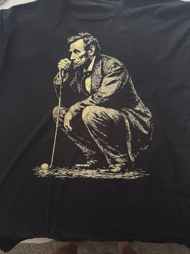 Customer's black Lincoln Reading The Green 5 T-Shirt laid flat, showing the cream graphic of Lincoln golfing.