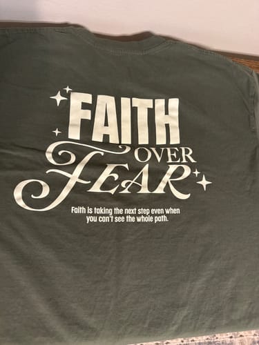 Customer's green COURAGE TEE laid flat, showing the 'FAITH OVER FEAR' graphic printed on the back.