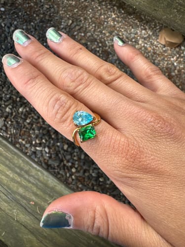 A customer's hand wearing the Navae Birthstone Ring, featuring a light blue pear-cut and a green radiant-cut stone.