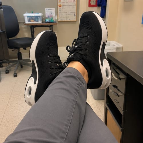 Reviewer relaxing with their feet up, wearing the black Ortho Walk 2.0™ shoes with thick white soles at work.