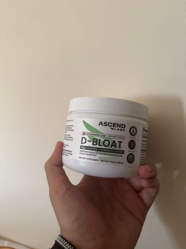 Reviewer's hand holding a white tub of D-BLOAT supplement powder against a plain background.