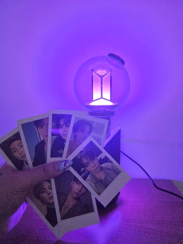Reviewer's BTS Official Light Stick Ver.4 on its cradle, glowing purple, with a hand holding photocards in the foreground.