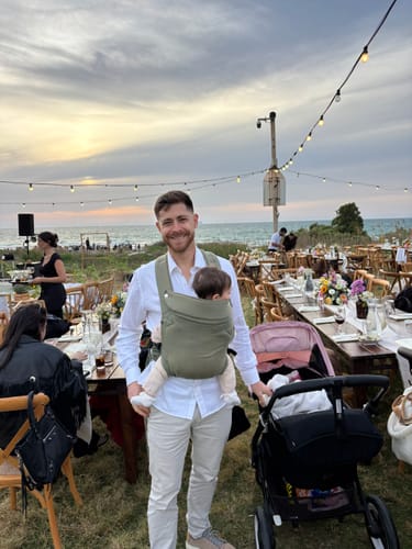 Reviewer standing outdoors while holding a baby in a muted green Urban Minimal baby Carrier.
