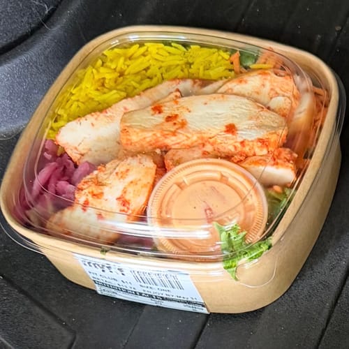 Customer's Harissa Chicken Bowl in its takeout container, showing sliced chicken, yellow rice, and dressing.