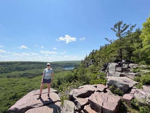 Reviewer wearing their 'I'd Rather Be On A Trail' T-Shirt while standing on a scenic, rocky overlook.