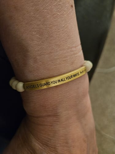 Customer's wrist wearing the 'ANGELS GUARD YOU IN ALL YOUR WAYS' bracelet, showing the engraved gold-colored bar and white beads.