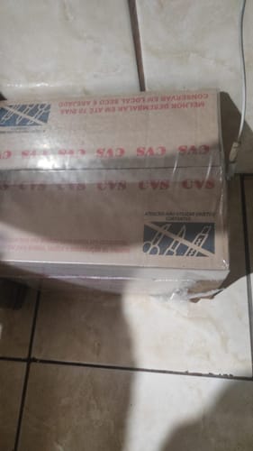 Cliente mostrando a caixa de papelão da Cesta Básica Jacarandá Plus, lacrada e em um piso de cerâmica.