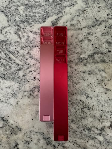 Customer's two shiny Weekly AM-PM Pill Cases, one pink and one red, resting side by side on a marble countertop.