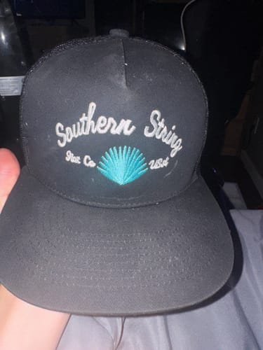 Customer holding a black trucker hat with 'Southern String' and a turquoise shell embroidered on the front.
