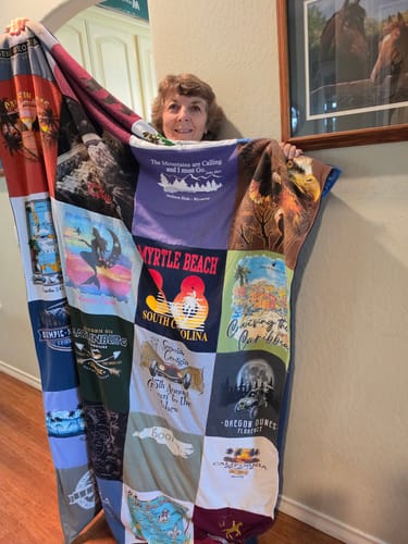 Reviewer holding up a large, colorful Project Repat T-Shirt Quilt made of many souvenir t-shirts.