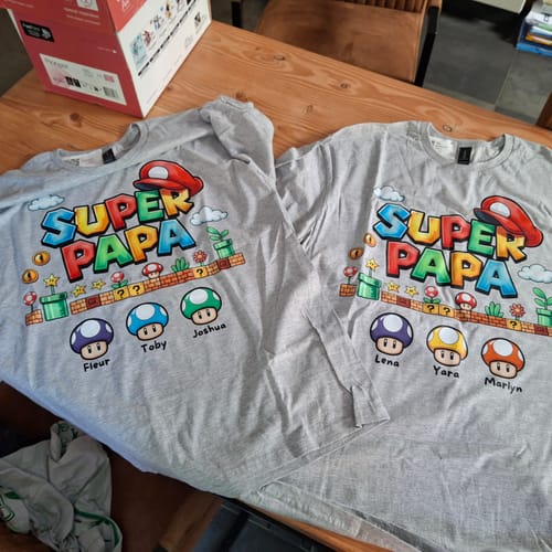 Kunde zeigt zwei personalisierte graue 'Super Papa'-Shirts, die auf einem Tisch liegen.