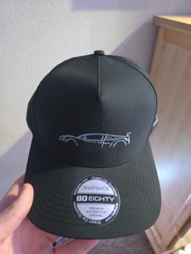 A person's hand holds the black 80Eighty DCG89 Hat, showing the front with its silver embroidered car graphic and branded sticker.