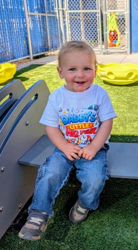 Customer's happy toddler sitting outdoors on a playground, wearing the 'Daddy's Little Helper' toddler tee.