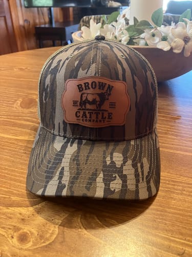 Reviewer's custom leather patch trucker hat in a green camo pattern, seen on a wooden table.