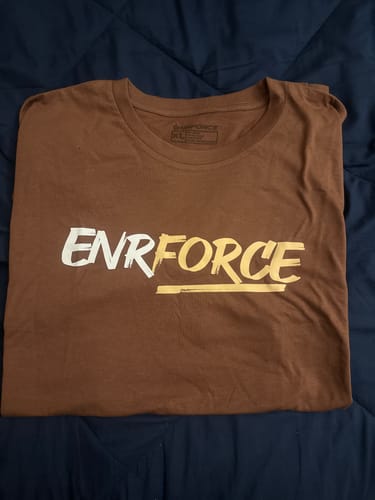 Reviewer's brown Mystery Tee with a white and yellow 'ENFORCE' graphic laid flat on a bed.