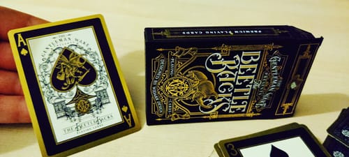 Customer holds the gold-bordered Ace of Spades from The Beetle Backs deck next to the matching black tuck box.