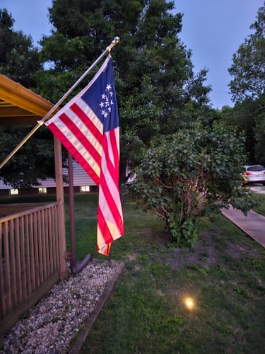 Customer's Betsy Ross Flag flying on a flagpole attached to a porch at dusk, illuminated from below.