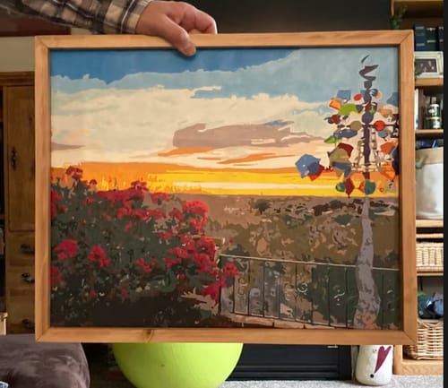 Customer holding the framed Custom Paint By Numbers artwork. The painting is a sunset landscape with red flowers in the foreground.
