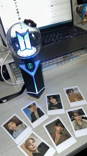 Customer's lit BTS Official Light Stick Ver.4 in its cradle, displayed on a desk with seven member photocards.