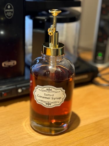 Nærbilde av en anmelders sirupdispenser med gullpumpe, fylt med mørk sirup og med en 'Salted Caramel Syrup'-etikett.
