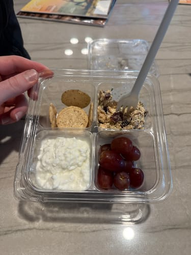 Customer holding the opened Protein Snack Pack, showing the chicken salad, cottage cheese, grapes, and crackers.