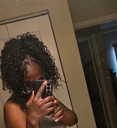 Customer's mirror selfie showcasing their new shoulder-length style with dark, curly Water Wave crochet hair.