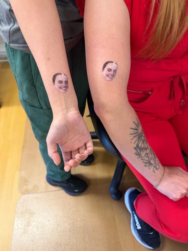 Two customers show off their matching Custom Photo Face Temporary Tattoos, one on a forearm and the other on an upper arm.