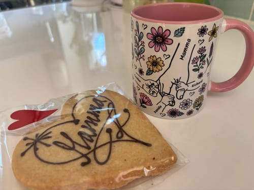 Kundens personliga Kärleksmugg med rosa detaljer och handmotiv, fotograferad bredvid en hjärtformad kaka.