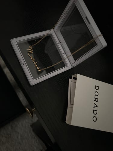 Customer's gold Mon Petit Name Necklace displayed in its open, clear case on a dark surface.