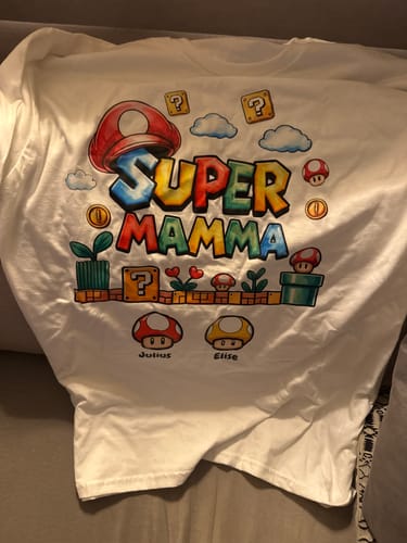 Rezensent zeigt das personalisierte weiße T-Shirt 'Super Mamma' mit Videospiel-Aufdruck und den Namen Julius und Elise.