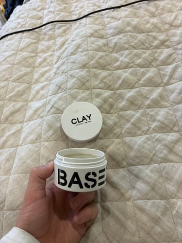Reviewer holding an open white jar of 'Base' hair product with the 'Clay' lid in the background on a quilt.