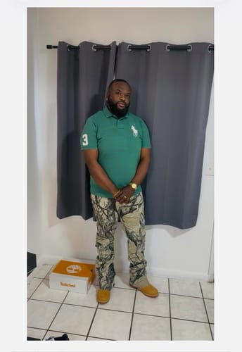 Customer wearing Vortex Cargo Stacked Denim in woodland camo, standing in front of a curtain.