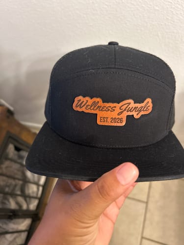 Customer holding a black seven-panel hat with a custom 'Wellness Jungle' engraved leather patch.