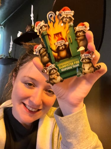 Customer smiling and holding up her personalized Raccoon Dumpster Fire Response Team ornament with custom names on the hats.