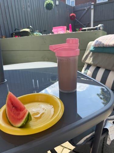 Customer's Slim Meal Replacement shake in a pink shaker on a sunny patio table with a slice of watermelon.