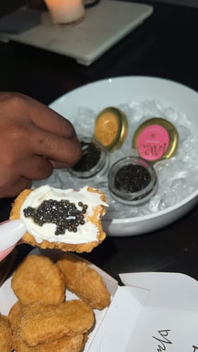 Customer's hand holds a cracker with cream cheese and caviar over a box of chicken nuggets, with jars of caviar on ice.