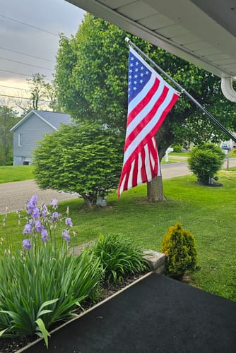 Customer's vibrant Ultra Heavy Duty American Flag displayed on a pole attached to their house, with a green yard in the background.
