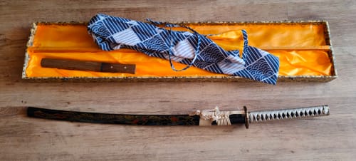 Reviewer's custom katana in its decorated black saya, next to its open presentation box and a cloth bag.