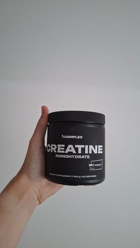 Customer's hand holding a black tub of Complex Creatine Monohydrate against a plain background.