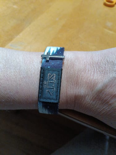 Customer's wrist showing the silver Cross Bracelet Charm attached to a patterned fabric bracelet against a wooden surface.