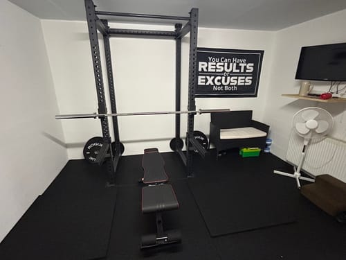 Rezensent zeigt das schwarze R8 Base 1 Power Rack in einem Heim-Fitnessstudio mit einer Hantelbank und einer Langhantel.