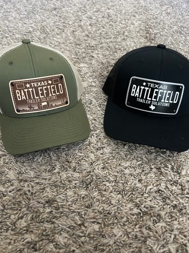 Customer's two hats, one green and one black, with custom Texas Engraved License Plate patches shown side-by-side on carpet.