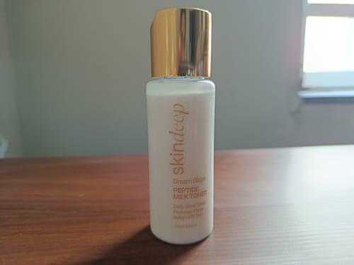 Customer's DREAM GLAZE - Peptide Milk Toner in a white bottle with a gold cap, sitting on a wooden desk.