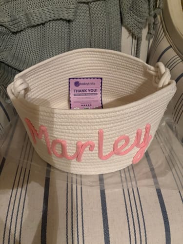 Customer's personalized white rope basket with the name 'Marley' in pink script, sitting on a striped chair.