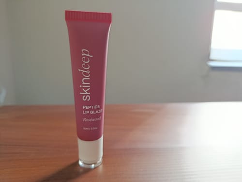 Customer's pink tube of Peptide Lip Glaze in the shade Rosewood standing on a wooden table.