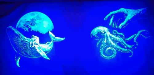 Customer's glowing Lunar Drifter shirt laid out next to another glowing shirt with an octopus and hand design.